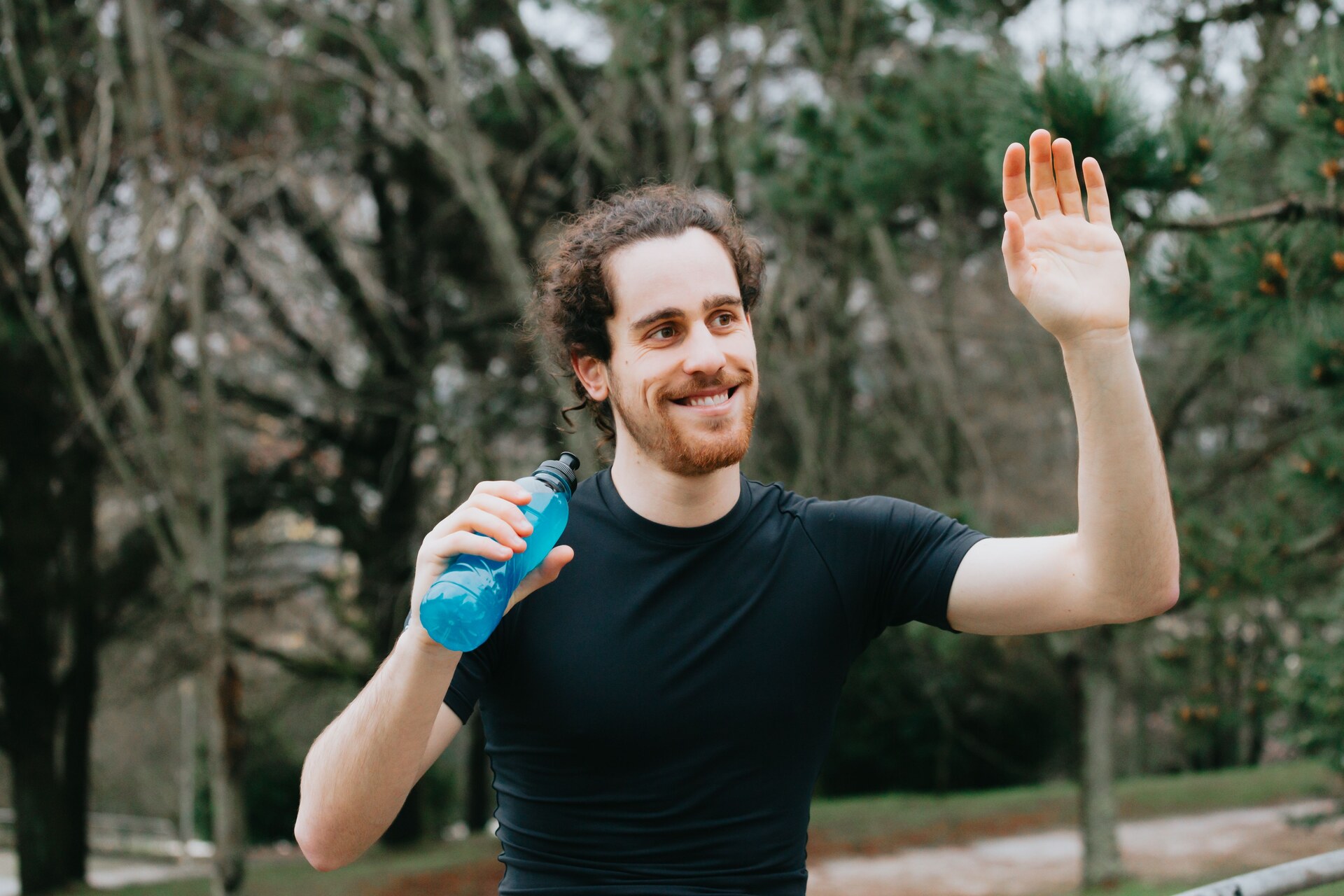 Smiling man waving outdoors in a park, holding a water bottle.